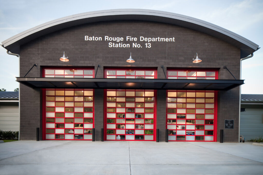 A modern fire station building with four large, red-framed glass garage doors, labeled "Baton Rouge Fire Department Station No. 13." Designed by leading Baton Rouge architects, the structure has a dark grey facade and exterior lighting above the garage doors. The surrounding area is clean and well-lit.