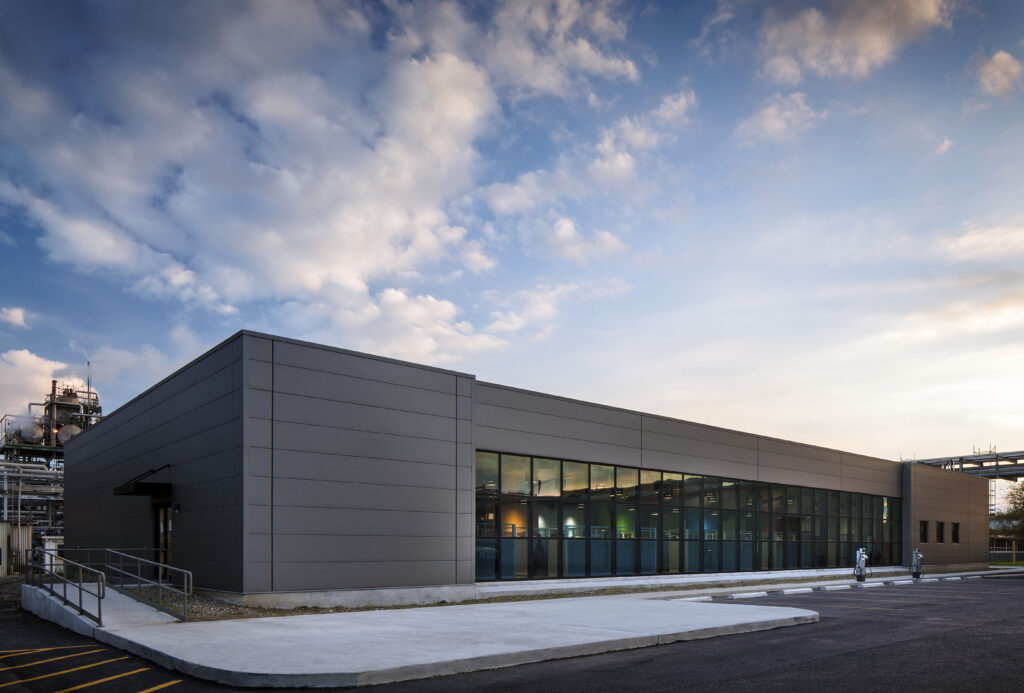 A modern industrial building with large windows stands against a backdrop of a partly cloudy sky. The building features clean lines and a flat roof. A concrete ramp leads to an entrance on the left side. The surrounding area is paved with asphalt.